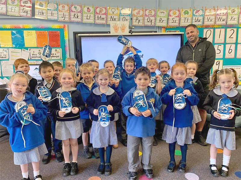 A classroom with young students holding up shoes, standing in front of a teacher and digital whiteboard.