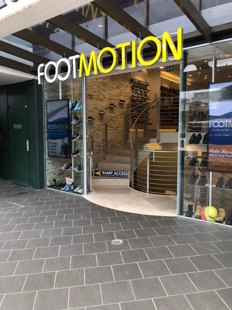 Entrance to FootMotion footwear and podiatry store, showing a staircase inside, shoe displays on the wall, and a ramp access sign.