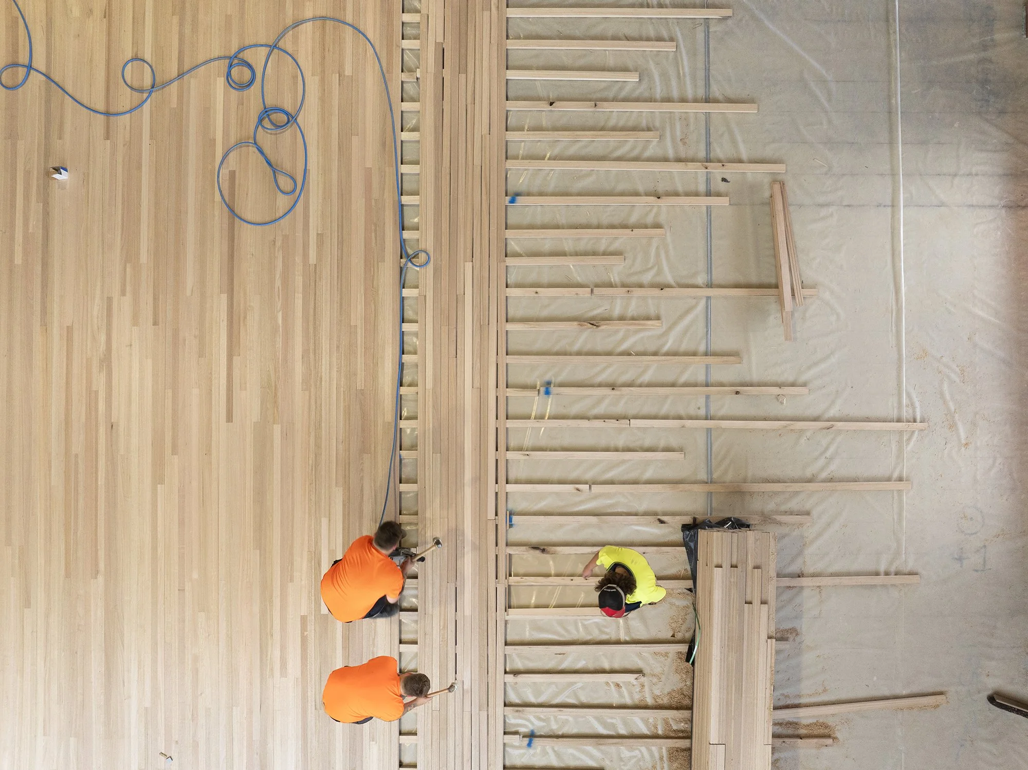 Two workers in orange shirts and one worker in yellow shirt installing wooden framing on a wall in a building under construction. The workers are kneeling and appear to be working on the lower part of the wall. The floor is light-colored hardwood, and the wall framing is made of light-colored wood with some horizontal supports. An extension cord is placed on the floor.