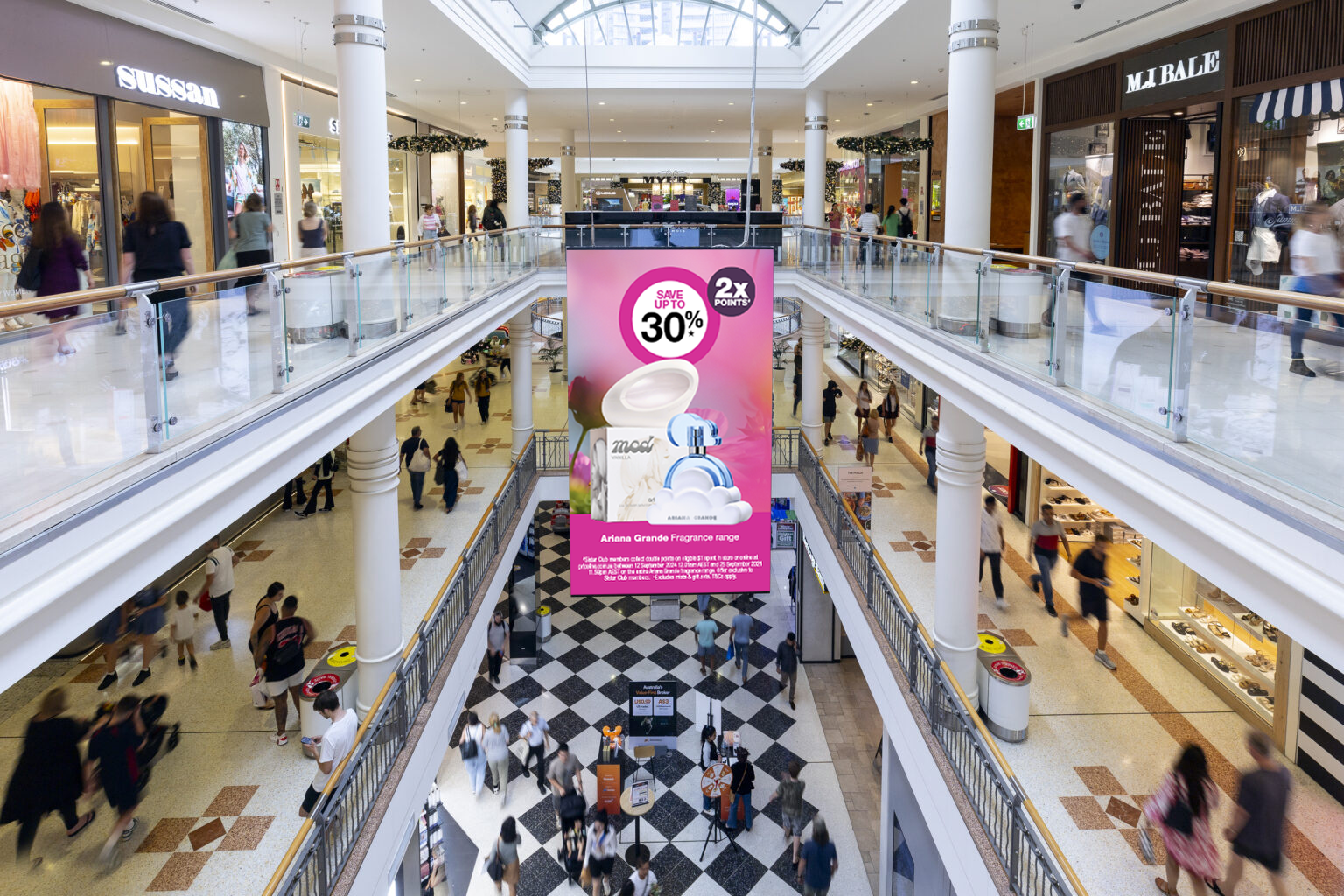 Interior of a multi-level shopping mall with various stores and shoppers walking on both floors. A large pink advertisement banner for Ariana Grande fragrances hangs from the ceiling.
