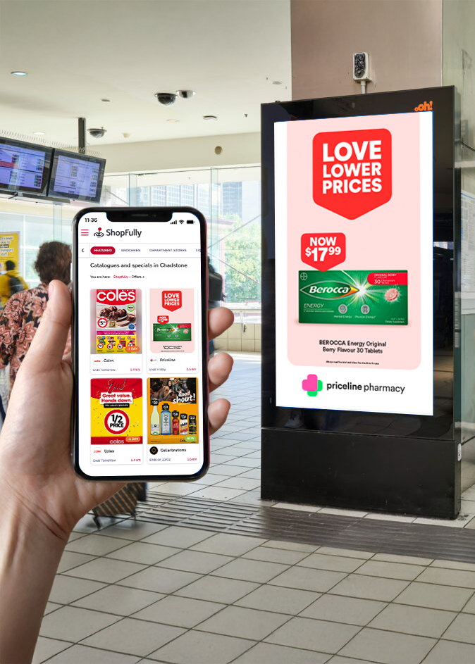 Person holding a smartphone displaying the ShopFully app near a digital billboard showing a sale on Berocca energy drink tablets at Priceline Pharmacy in a mall.