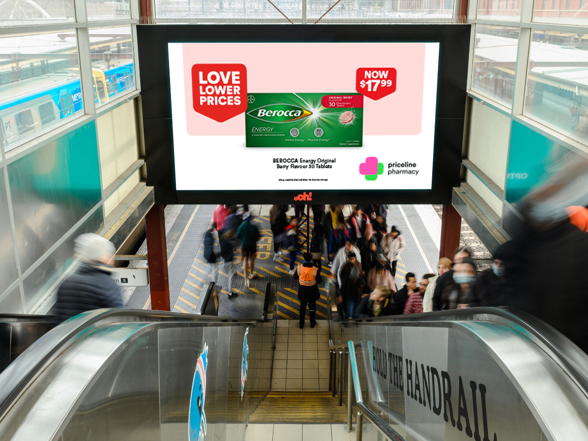 An escalator descending to a busy subway or train station entrance with a large digital advertisement screen above. The ad promotes Berocca energy supplements with a price of $17.99, featuring a green box of the product and logos for LoveLowerPrices and Priceline Pharmacy. Passengers are visible at the bottom of the escalator, walking through the station entrance.