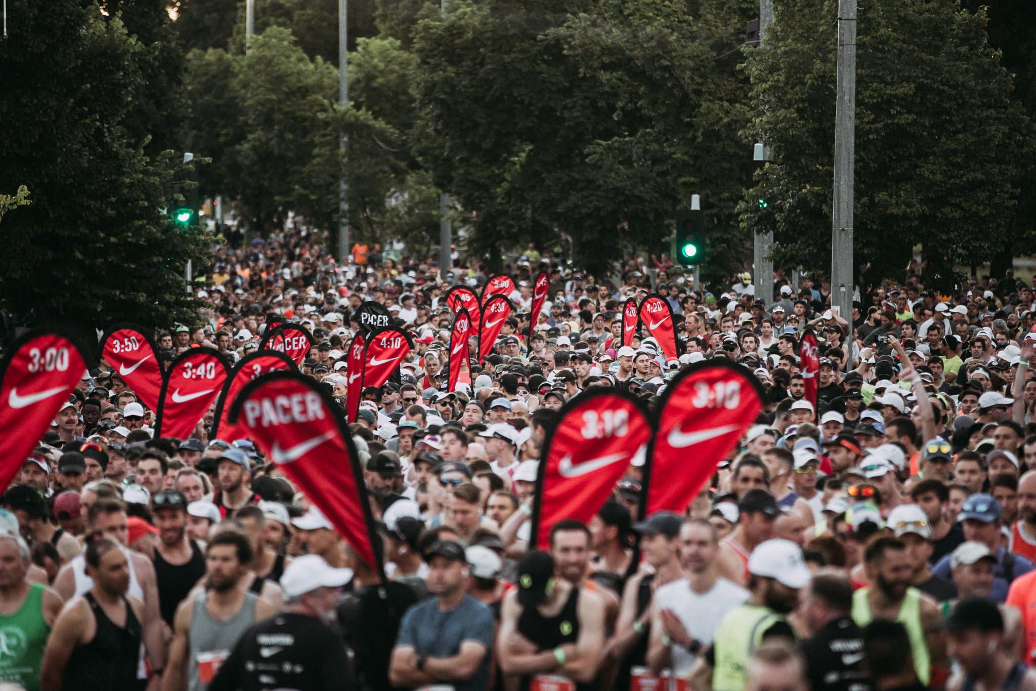 A large crowd of people participating in a marathon or running event on a city street, with red flags displaying Nike logos and pacing time markers, surrounded by trees and traffic signals.