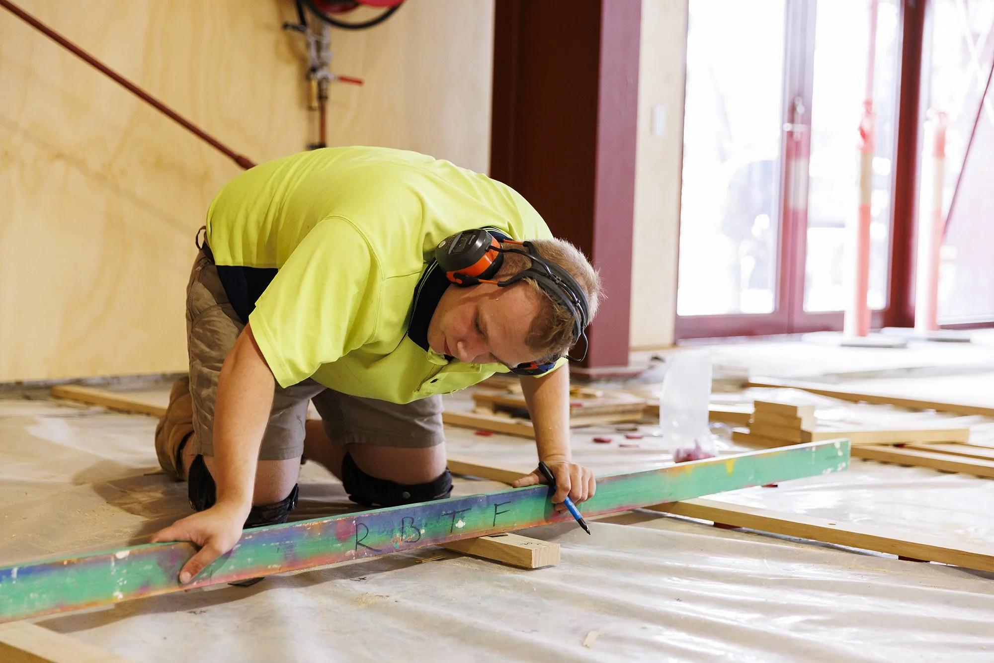 A man kneeling on the floor marking a wooden plank with a marker in a construction site.