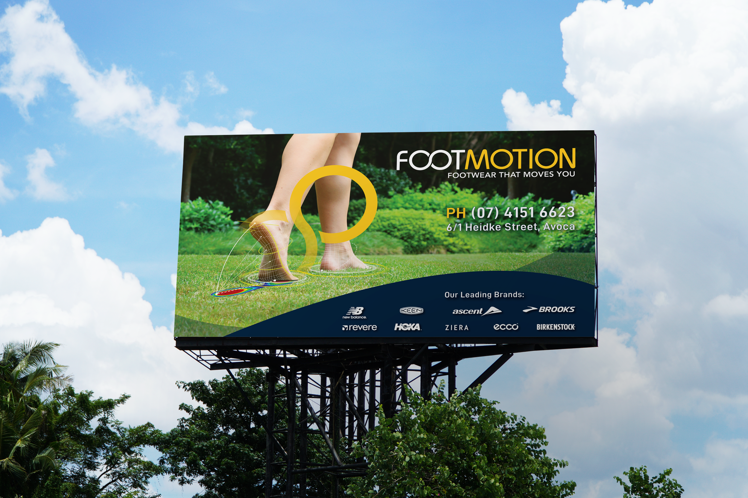 Billboard advertising FootMotion footwear store. The billboard features a person barefoot on grass, with an animation of digital footprints. Contact information and brand logos are displayed at the bottom. The background shows a blue sky with clouds and trees.