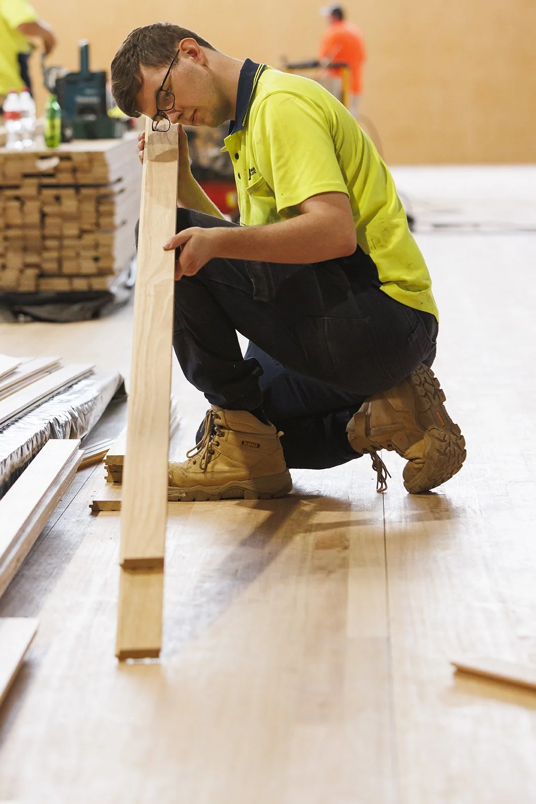 A man wearing glasses and a yellow shirt is kneeling on a wooden floor, measuring a long piece of wood in a workshop with other woodworking materials and workers in the background.