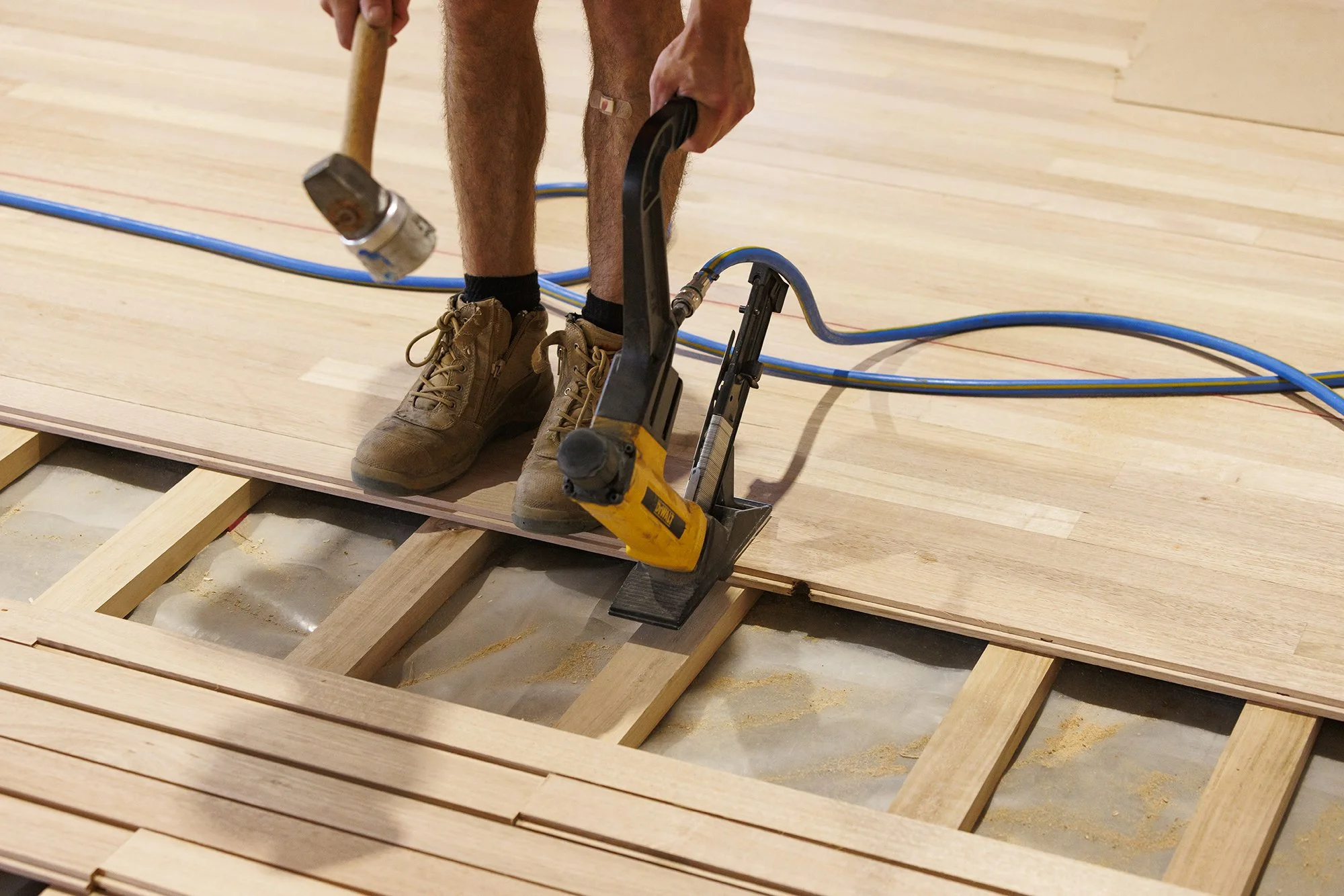 A person wearing work boots uses a nail gun to attach hardwood flooring to wooden subflooring in a woodworking project.