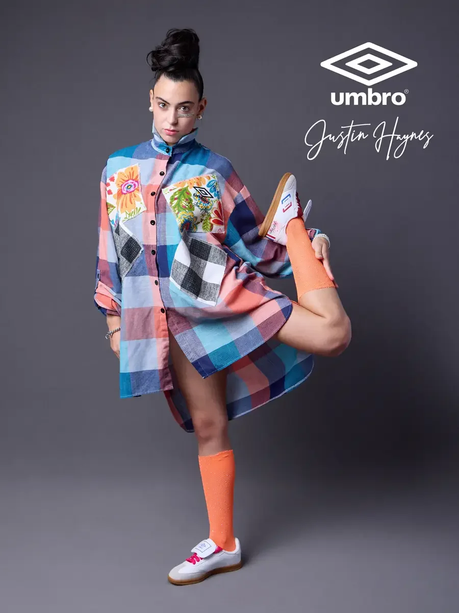 Lookbook photography for the Umbro France collection. Vanessa holding one leg raised with hand gripping the sneaker in an oversized multicolor plaid shirt dress with floral patchwork panels and orange football knee socks, styled by Justin Haynes, mod