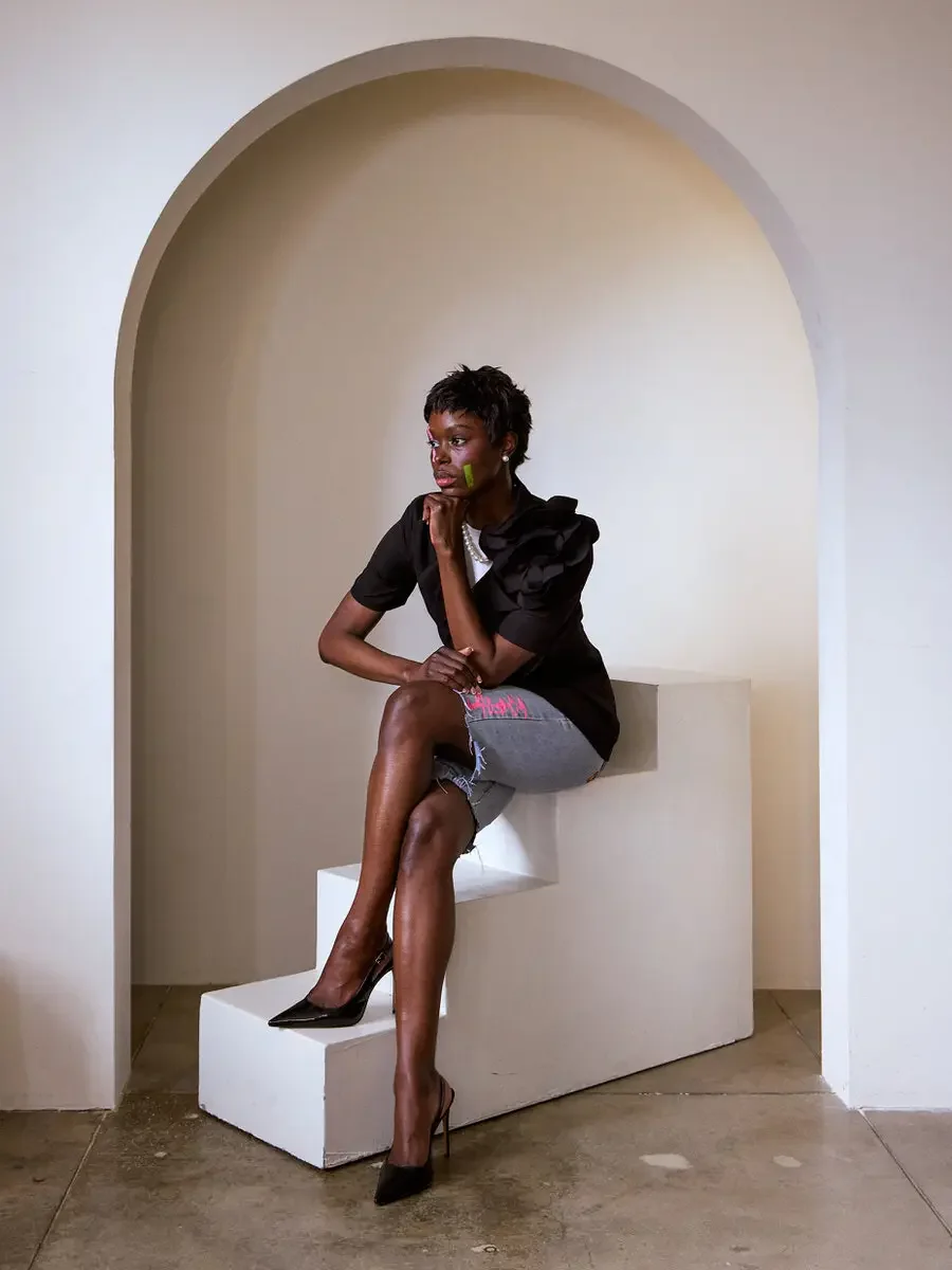 JUS10H Black Summer Jacket content library, female model in black blazer with oversized floral corsage, seated on white steps inside arch looking away, denim shorts with JUS10H logo, black slingback heels, warm neutral background, photographed by Sco