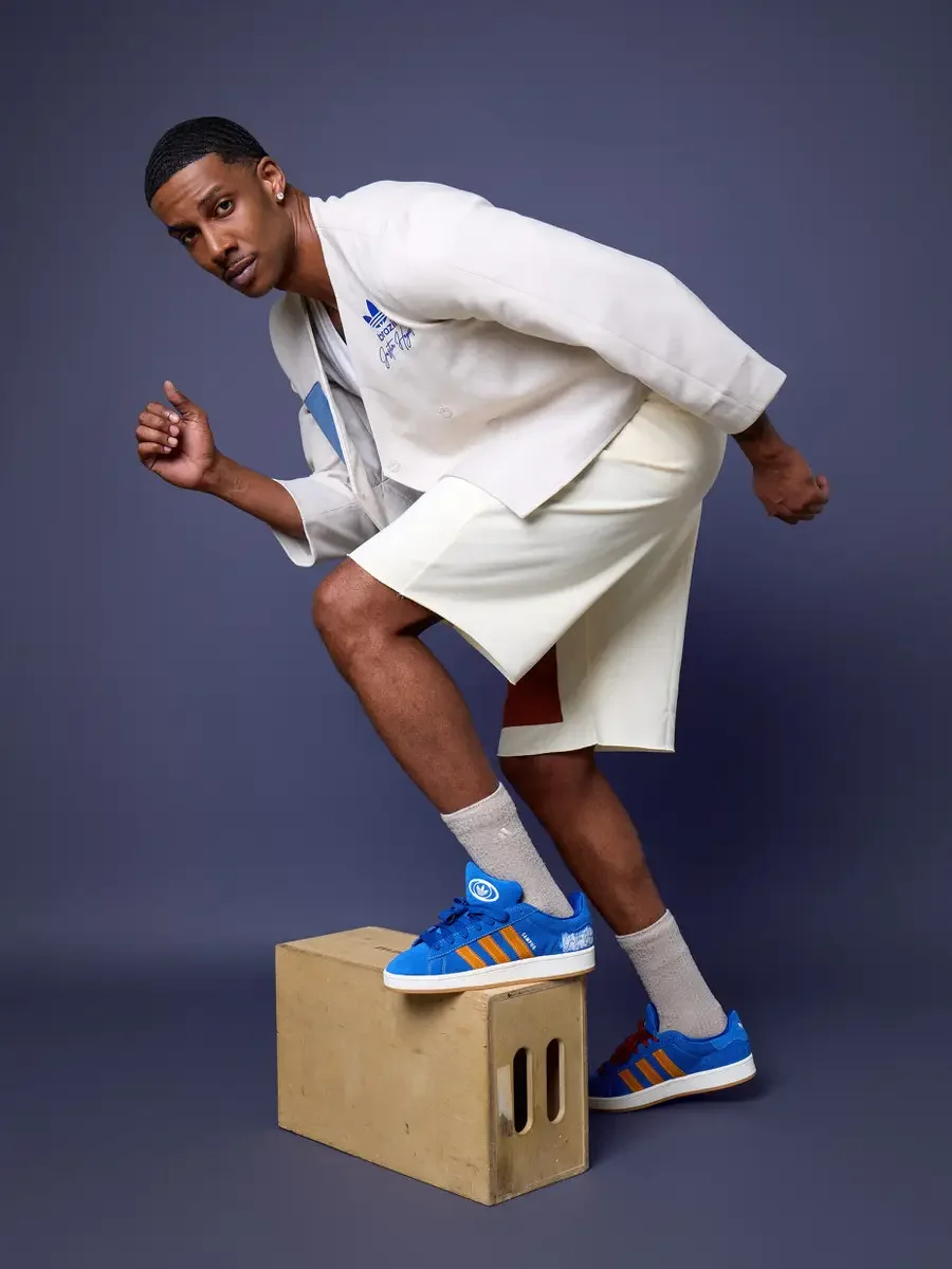 JUS10H x Adidas Brasil campaign, model leaning forward with one foot on wood block, white patchwork open robe jacket with blue and terracotta suede panels over cream patchwork shorts, Adidas Campus 00s in royal blue suede with orange stripes, periwin