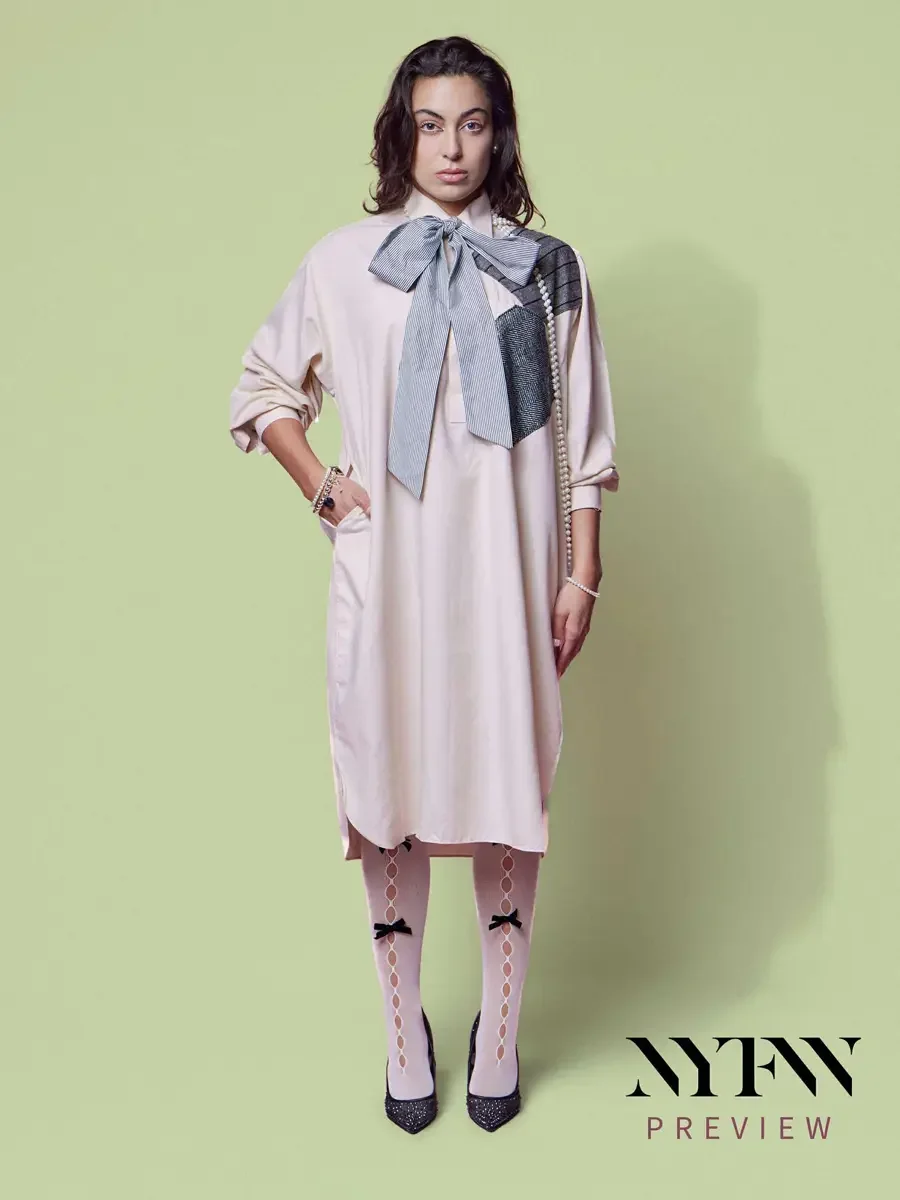 JUS10H 2025 billboard Times Square, model in blush oversized shirt-dress with striped bow, metallic and herringbone patchwork panel, long pearl necklace, bow-detail sheer tights and glitter pumps, hand on hip, sage green background, photographed by S