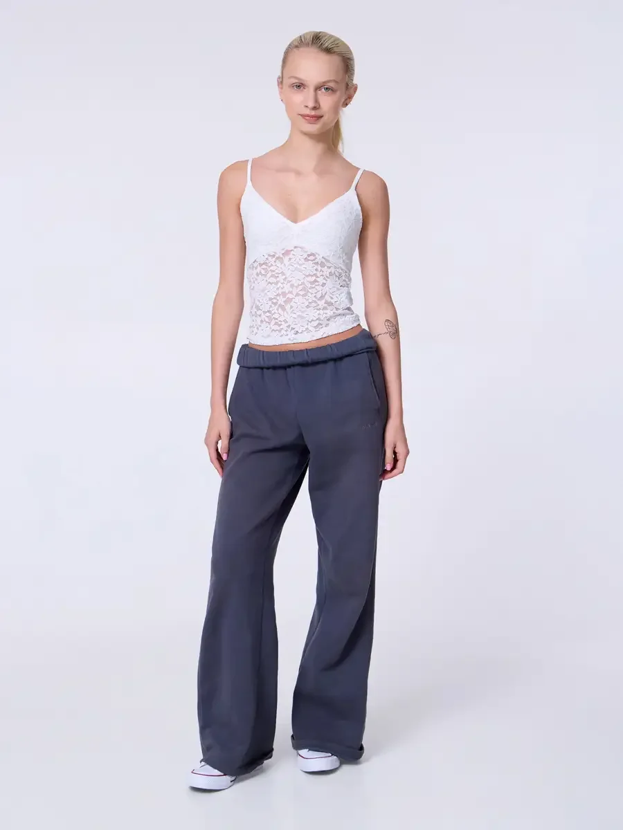 Model portfolio development session, commercial look, white lace crop camisole top and wide-leg charcoal sweatpants with elastic waistband, full-length, facing camera, arms relaxed at sides, neutral expression, white Converse sneakers, light gray bac