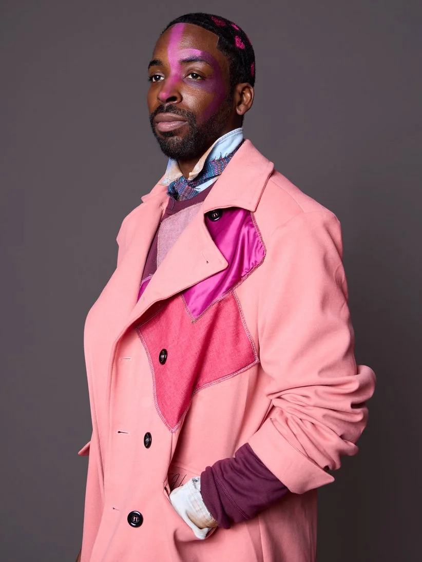 A man with a beard wearing a pink coat over different layered clothing, with pink and purple makeup and a textured hairstyle, standing against a neutral background.