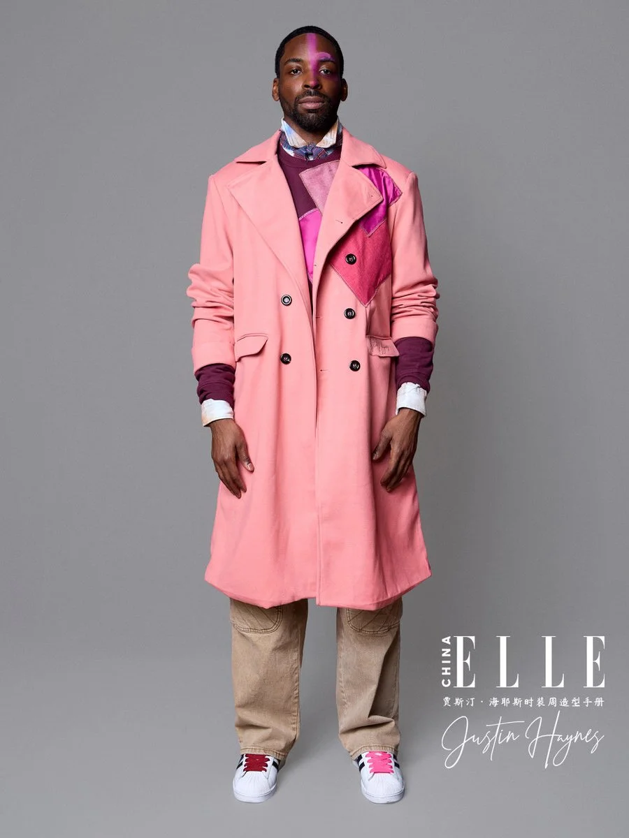 A man standing against a gray background wearing a pink trench coat over layered clothing, including a maroon sweater and shirt, beige pants, and white sneakers with pink and black accents, with Chinese text and the ELLE magazine logo in the bottom r