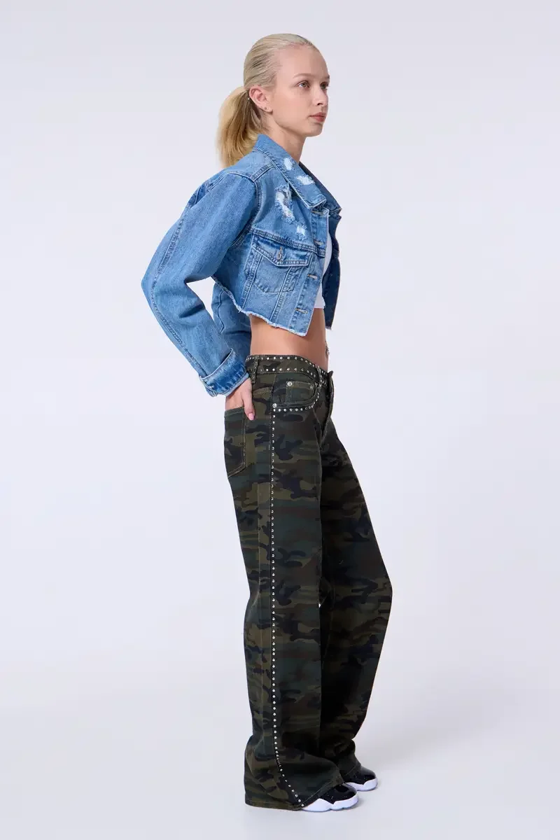 Model portfolio development session, commercial look, cropped distressed blue denim jacket over white crop tank, low-rise studded camouflage wide-leg trousers with silver stud trim at waistband and side seam, full-length, profile, one hand on hip, ga