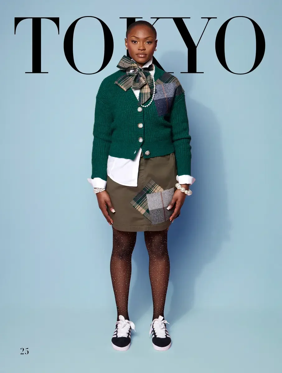 JUS10H 2025 Tokyo lookbook, forest green ribbed cardigan with tartan patch and bow over white shirt and olive mini skirt with tartan patch and fishnet knee socks with Adidas sneakers, light blue background, photographed by Scott Parker Photo for the 