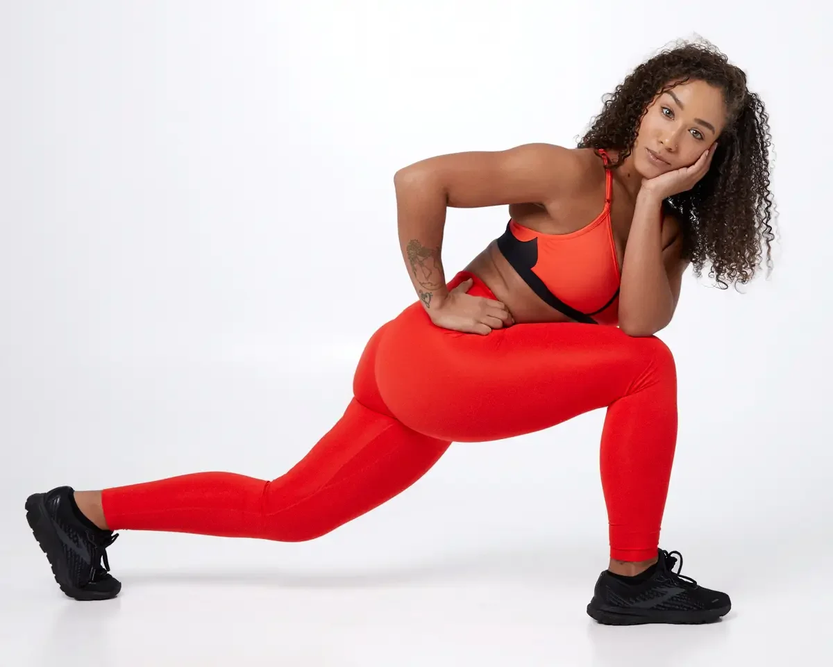 Model portfolio development session, commercial athletic look, orange sports bra with black underband, high-waist orange leggings, black athletic sneakers, full-length, low lunge position, hand to cheek, relaxed expression, white background