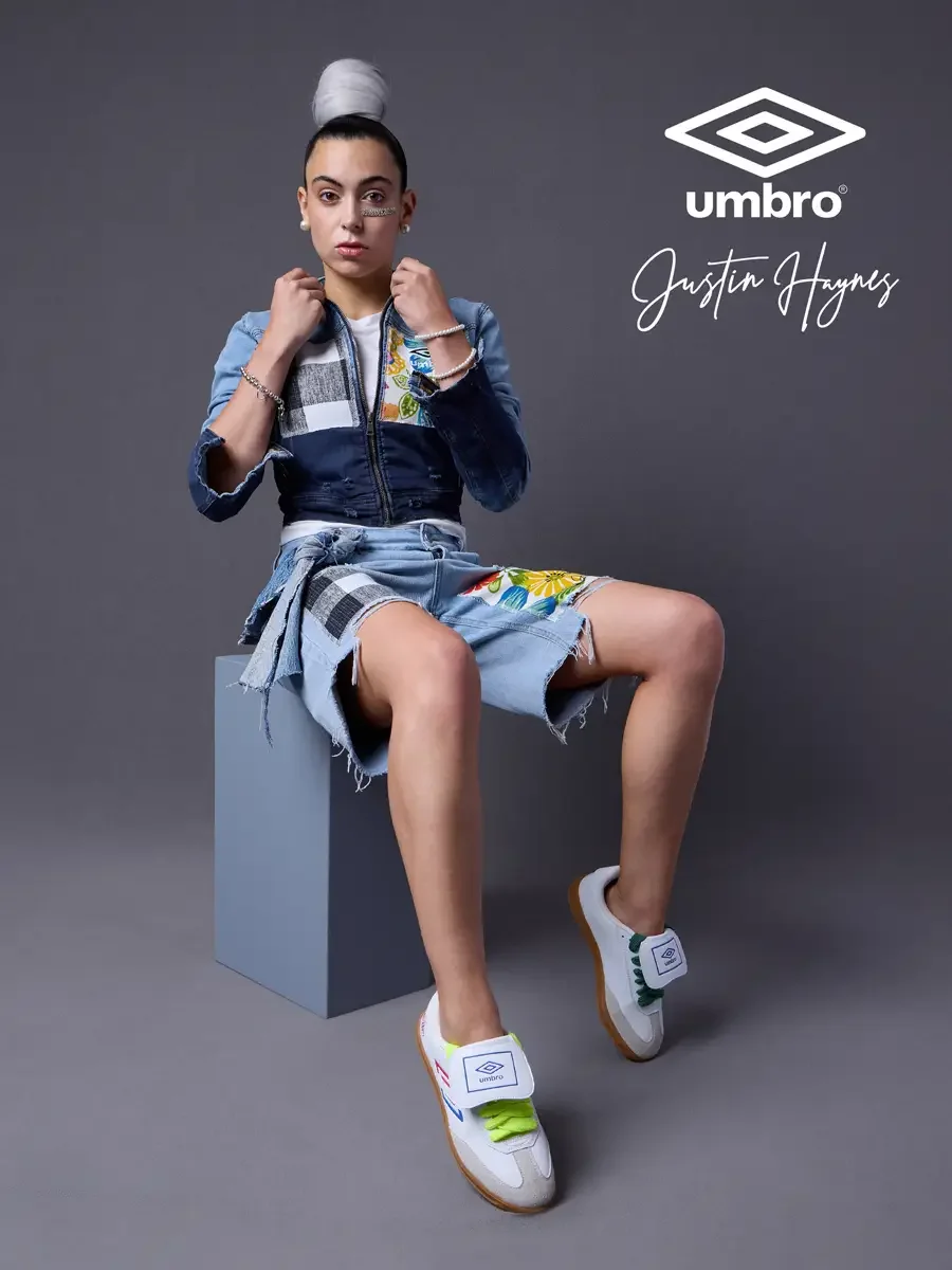 Elevated soccer style for the Umbro France collection. Vanessa seated pulling her jacket collar with both hands in a cropped two-tone denim moto jacket with floral patchwork panels and matching distressed denim bermuda shorts, styled by Justin Haynes