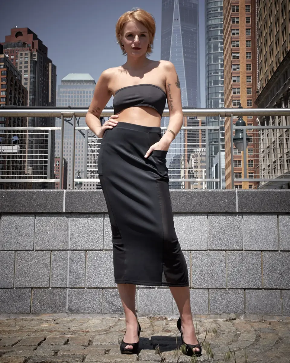 Satine, full length, hand on hip, black strapless bandeau crop top, black satin fitted midi skirt with wide waistband and front slit hem, black satin peep-toe stiletto heels, script tattoo on right arm visible, Battery Park esplanade, One World Trade
