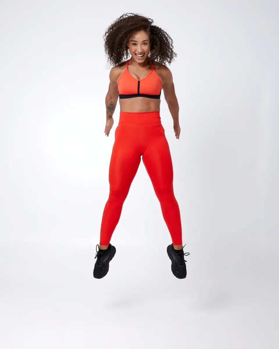 Model portfolio development session, commercial athletic look, orange sports bra with black underband, high-waist orange leggings, black athletic sneakers, full-length, mid-jump, smiling, white background