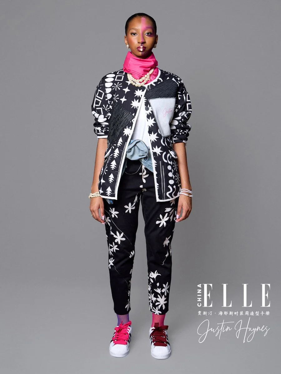 A woman in a black and white patterned jacket and pants with floral designs, pink sneakers, a pink neck scarf, and pearl jewelry poses against a plain gray background for Elle China magazine, with text mentioning Justin Hayes.