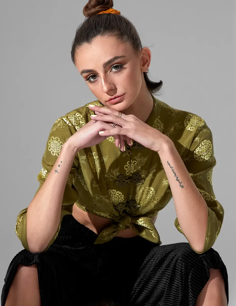 Macy, close crop, seated, hands together at chin, olive green satin jacquard tied crop top with gold medallion print and frog closures, amber scrunchie, script tattoo and star tattoos on forearm visible, New York studio test shoot, photographed by Sc