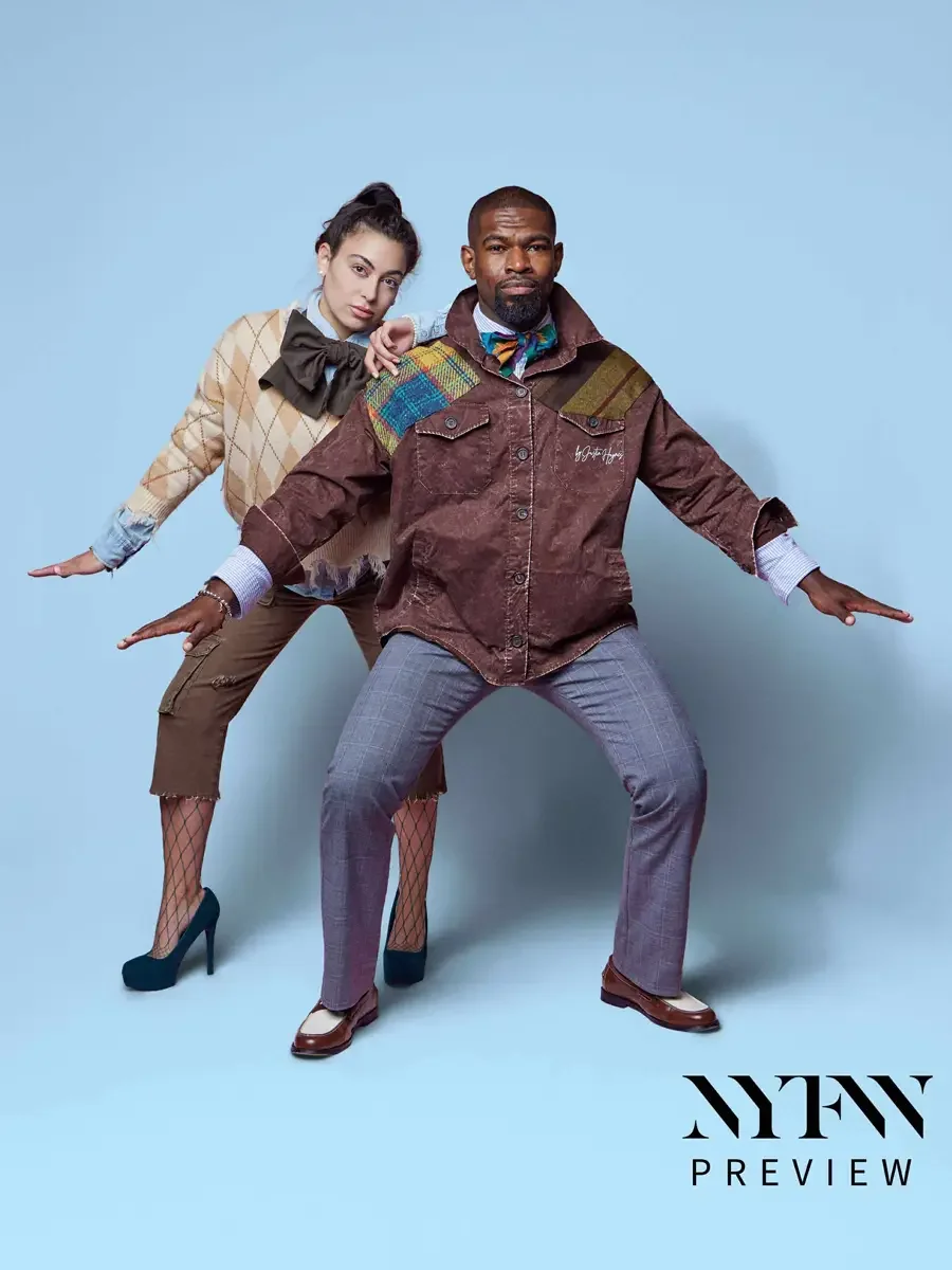 JUS10H 2025 billboard Times Square, two models mid-movement, female in camel argyle fringe sweater with multicolor tartan patchwork panel, olive bow, and brown cargo capris over fishnet tights, male in brown washed field jacket with multicolor tartan