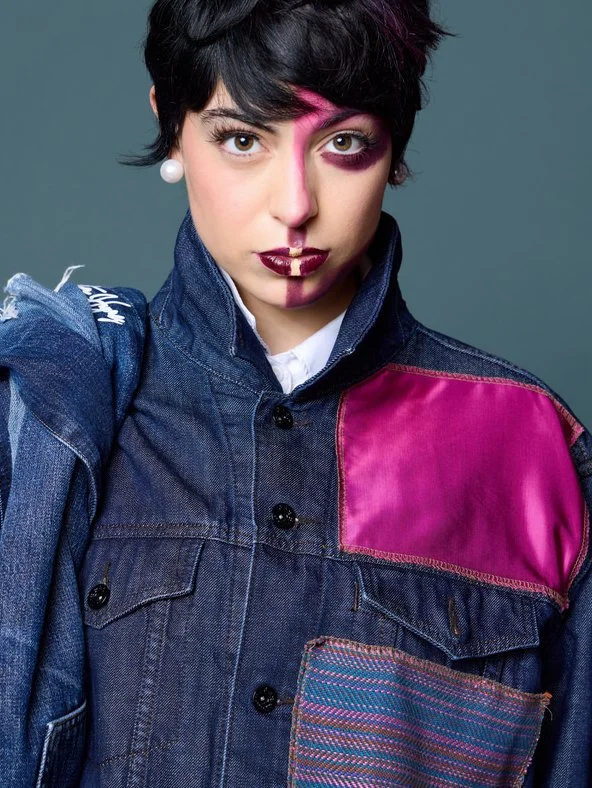 Portrait of a woman with short black hair, wearing a denim jacket with pink and patterned fabric accents, and bold makeup including face paint and lipstick.