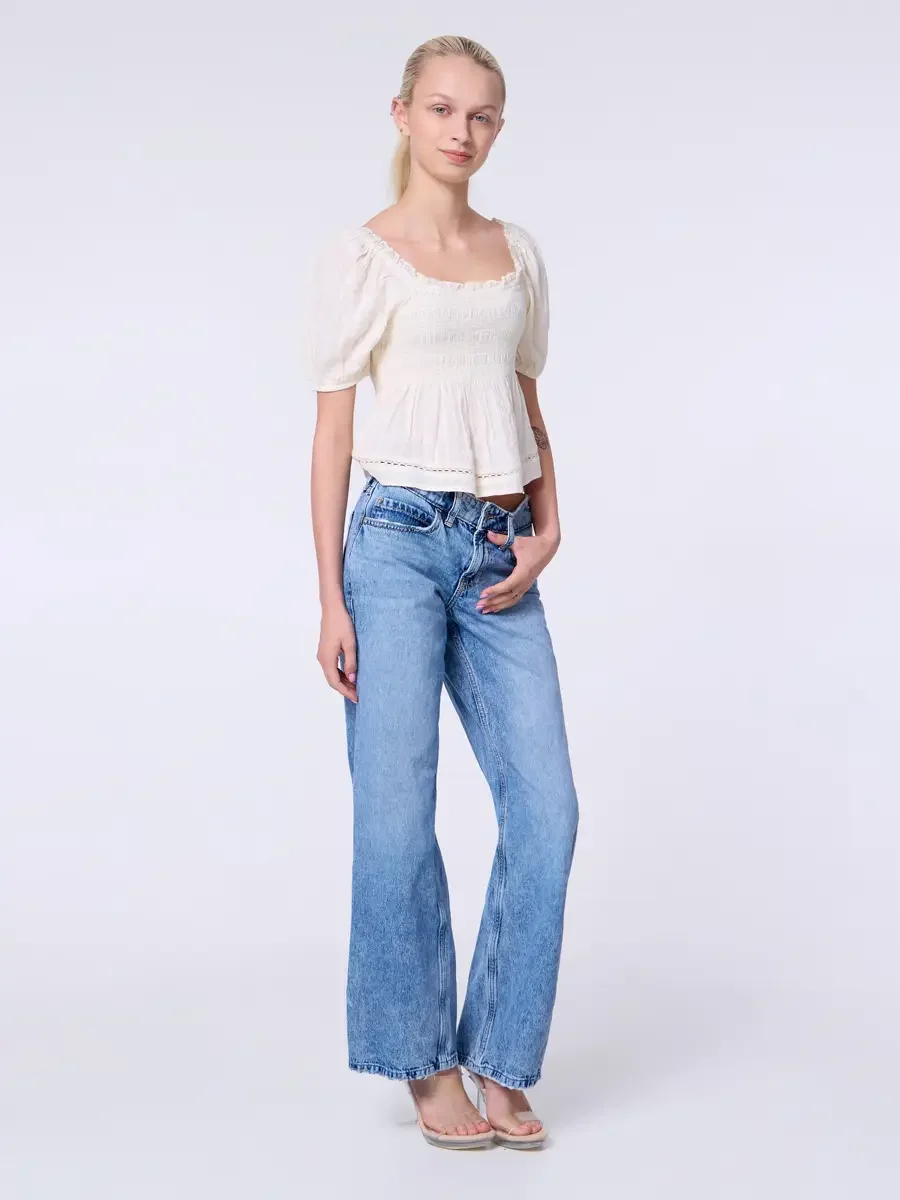 Model portfolio development session, commercial look, cream smocked square-neck puff-sleeve crop top and low-rise light wash wide-leg jeans, full-length, slight turn to camera, one hand in pocket, soft smile, puff sleeve silhouette and smocked bodice