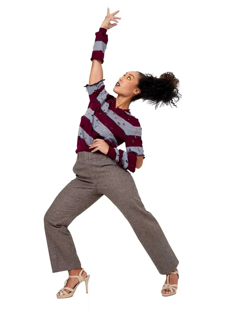 Model portfolio development session, high fashion editorial look, burgundy and heather grey destroyed-knit stripe sweater with raw-edge cutout sleeves, straight-leg heathered trousers, nude t-strap heels, blazer removed, full-length, one arm raised, 