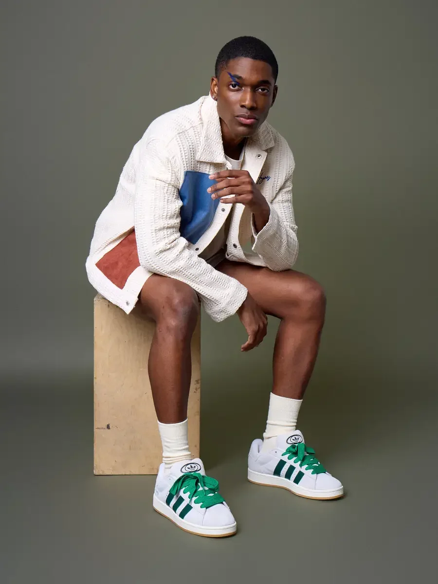 JUS10H x Adidas Brasil campaign, model seated on wood block with hands clasped, white textured patchwork jacket with blue and terracotta suede panels, Adidas Campus 00s in white with green stripes and green laces, sage green background, photographed 