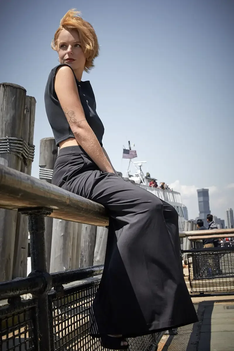 Satine, full length, seated on pier railing, gaze upward, black structured cap-sleeve crop top, black wide-leg pleated palazzo pants, floral tattoo on left arm visible, harbor and ferry in background, Battery Park pier test shoot, photographed by Sco