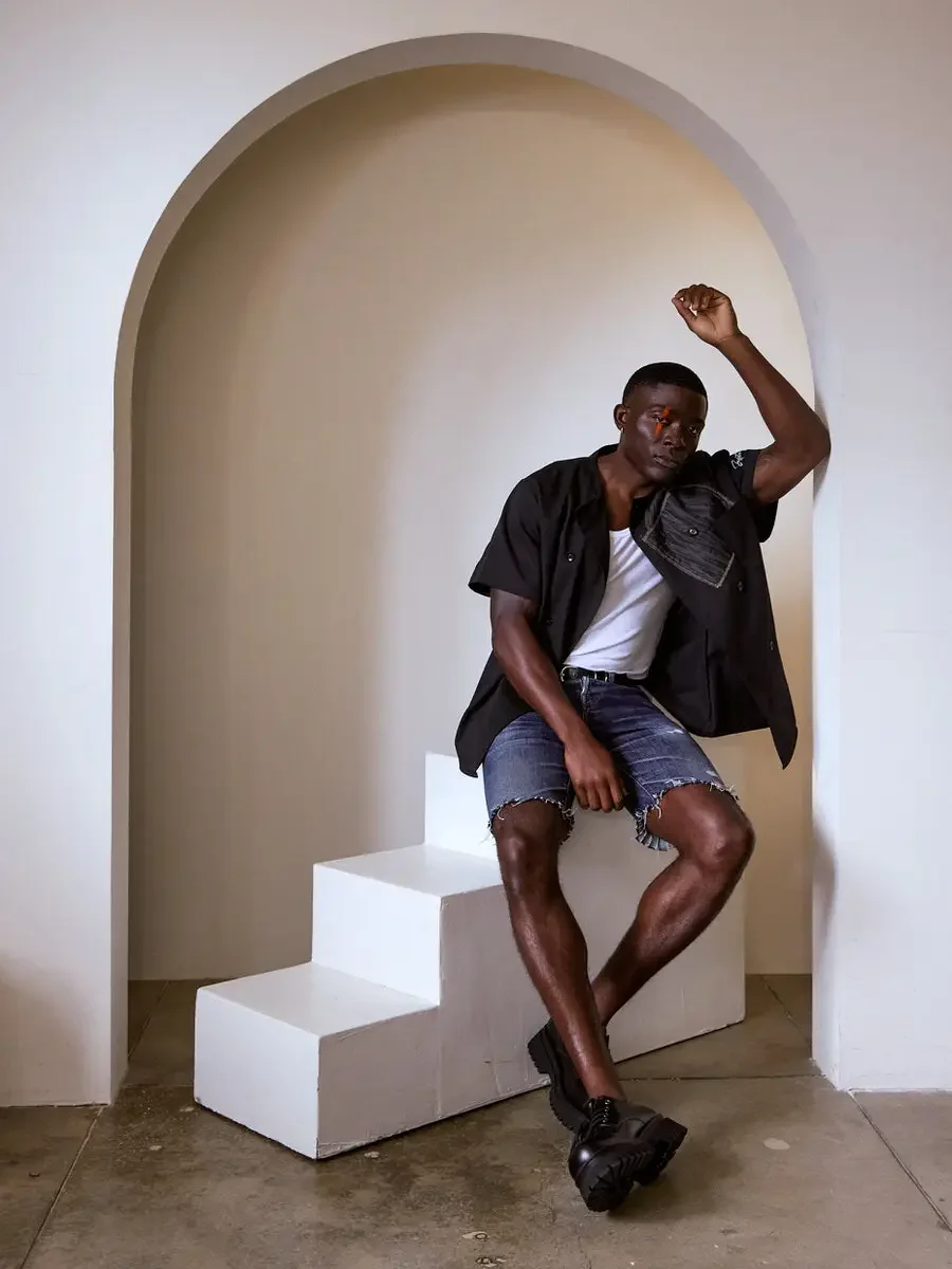 JUS10H Black Summer Jacket content library, male model in black double-breasted short-sleeve jacket with gray patchwork chest pocket, seated on white steps inside arch, warm neutral background, photographed by Scott Parker Photo