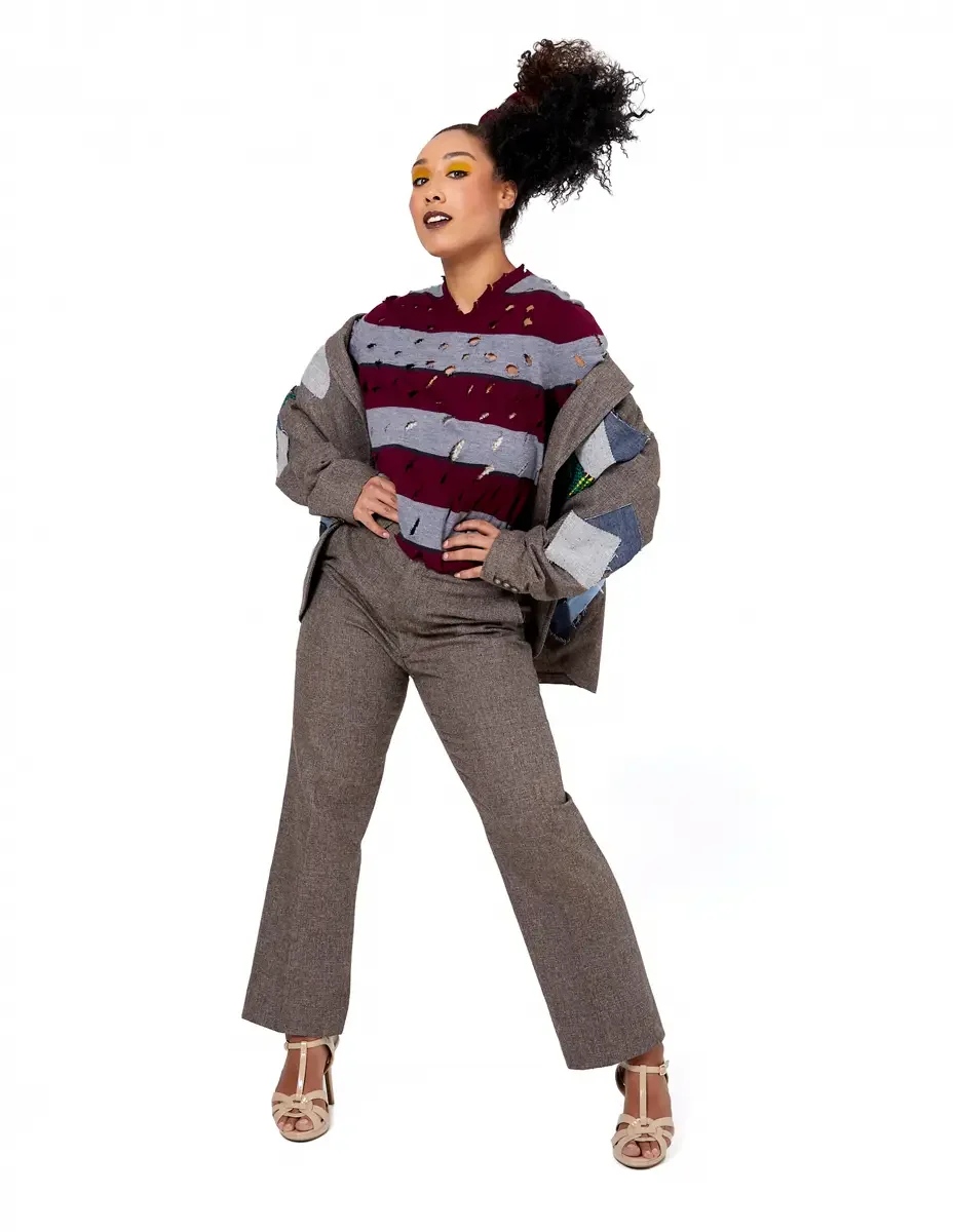 Model portfolio development session, high fashion editorial look, burgundy and heather grey destroyed-knit stripe sweater with raw-edge cutout sleeves, straight-leg heathered trousers, nude t-strap heels, blazer removed, full-length, hands at waist, 