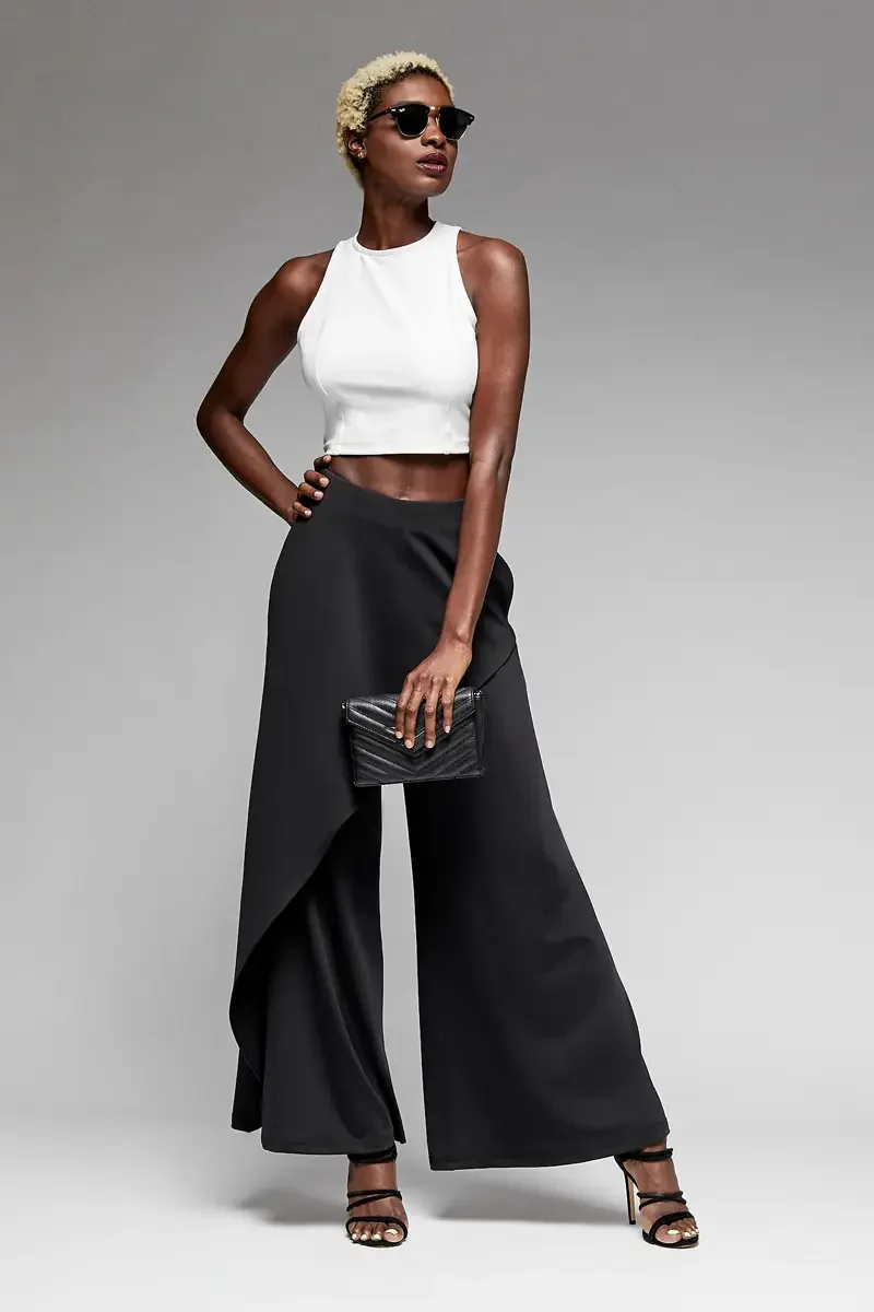 Minika Ko lookbook, black wide-leg trousers with asymmetric wrap skirt overlay and white halter crop top, three-quarter view, draped skirt panel falling asymmetrically over wide-leg silhouette, black quilted clutch, dark sunglasses, photographed by S
