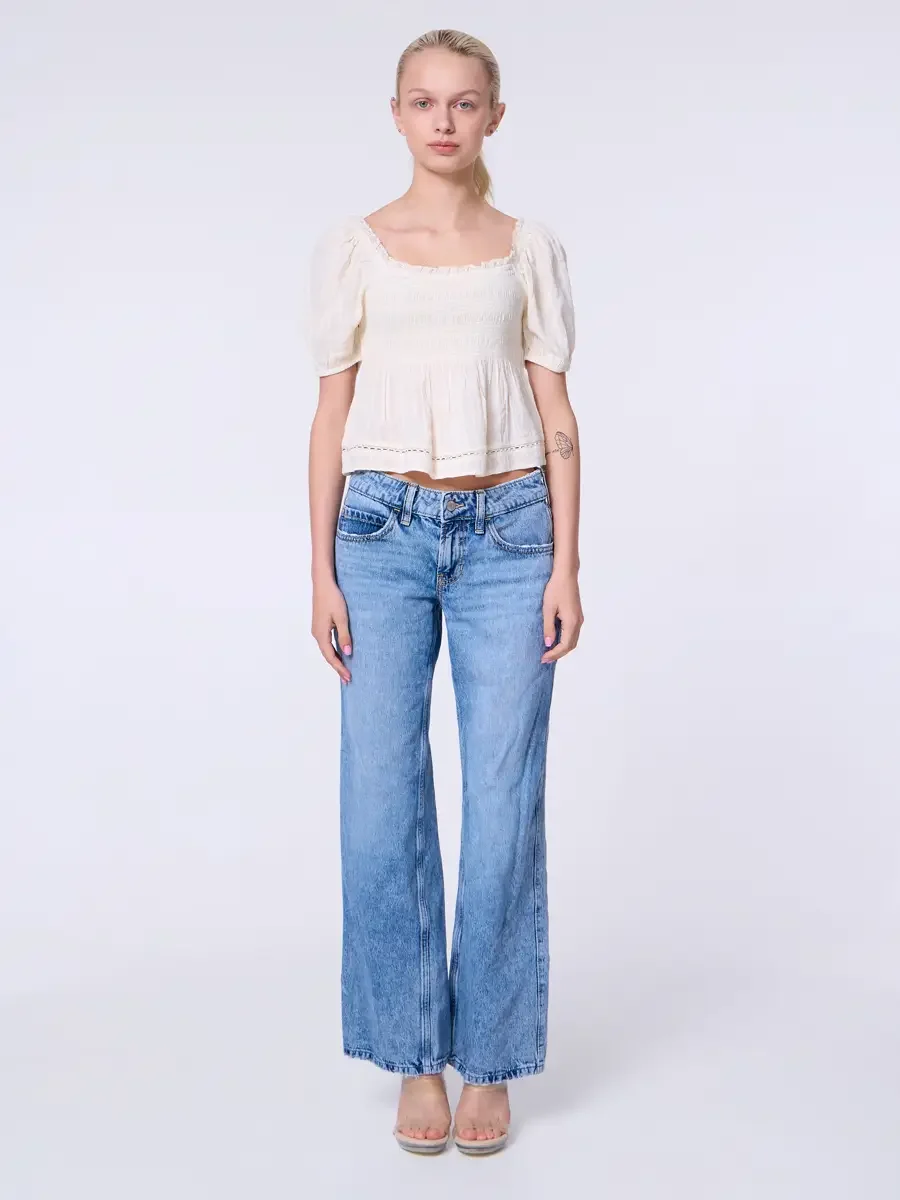 Model portfolio development session, commercial look, cream smocked square-neck puff-sleeve crop top and low-rise light wash wide-leg jeans, full-length, facing camera, arms relaxed at sides, neutral expression, crochet hem trim at top visible, nude 
