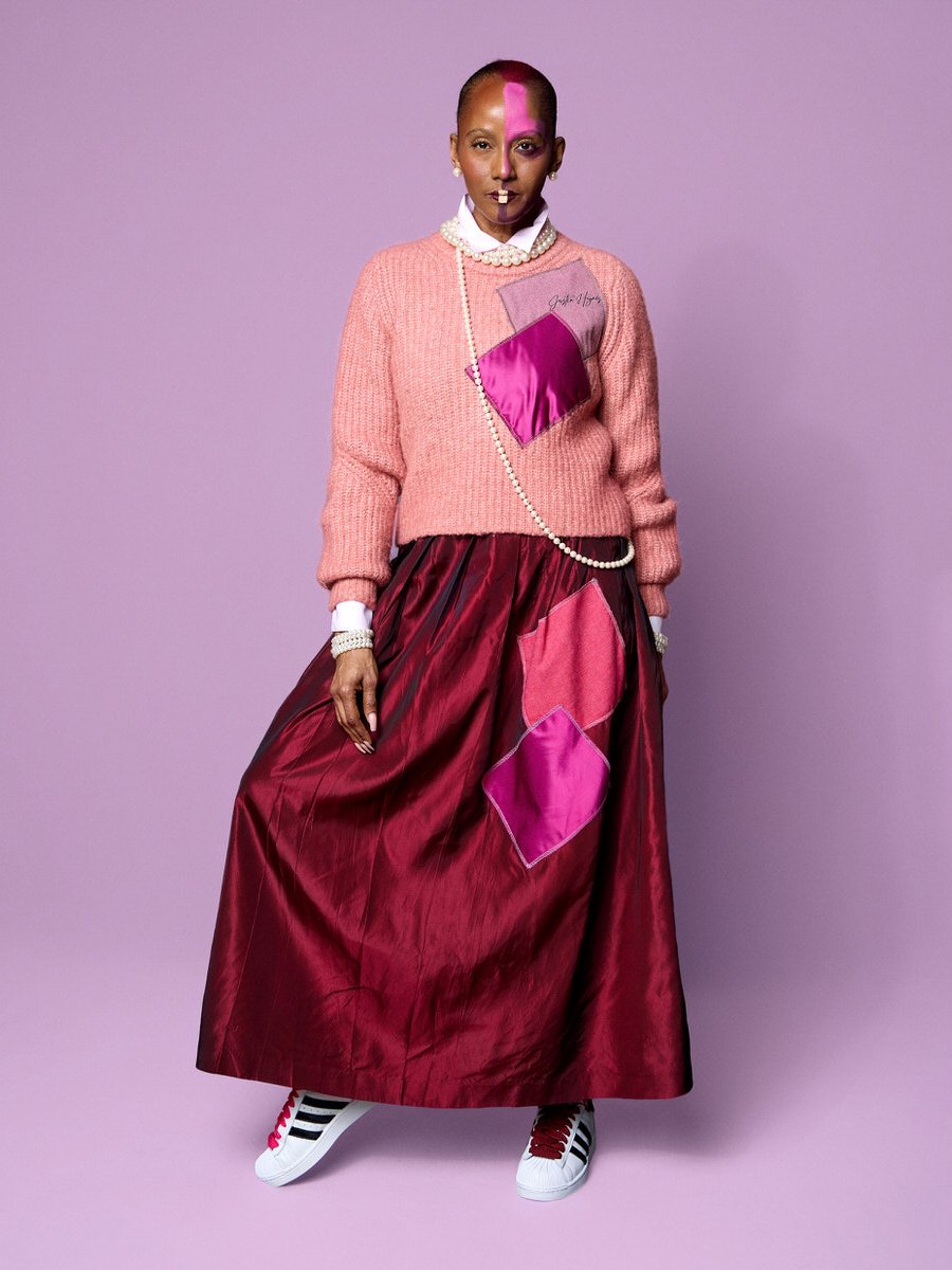 Woman wearing a pink sweater, pearl necklace, long burgundy satin skirt with pink patches, and white sneakers with black and red stripes, standing against a lavender background.