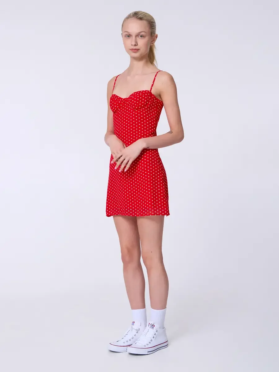 Model portfolio development session, commercial look, red polka dot sweetheart mini dress with spaghetti straps, full-length, three-quarter turn, hands loosely clasped at waist, composed expression, sweetheart bodice seaming visible in relief, white 