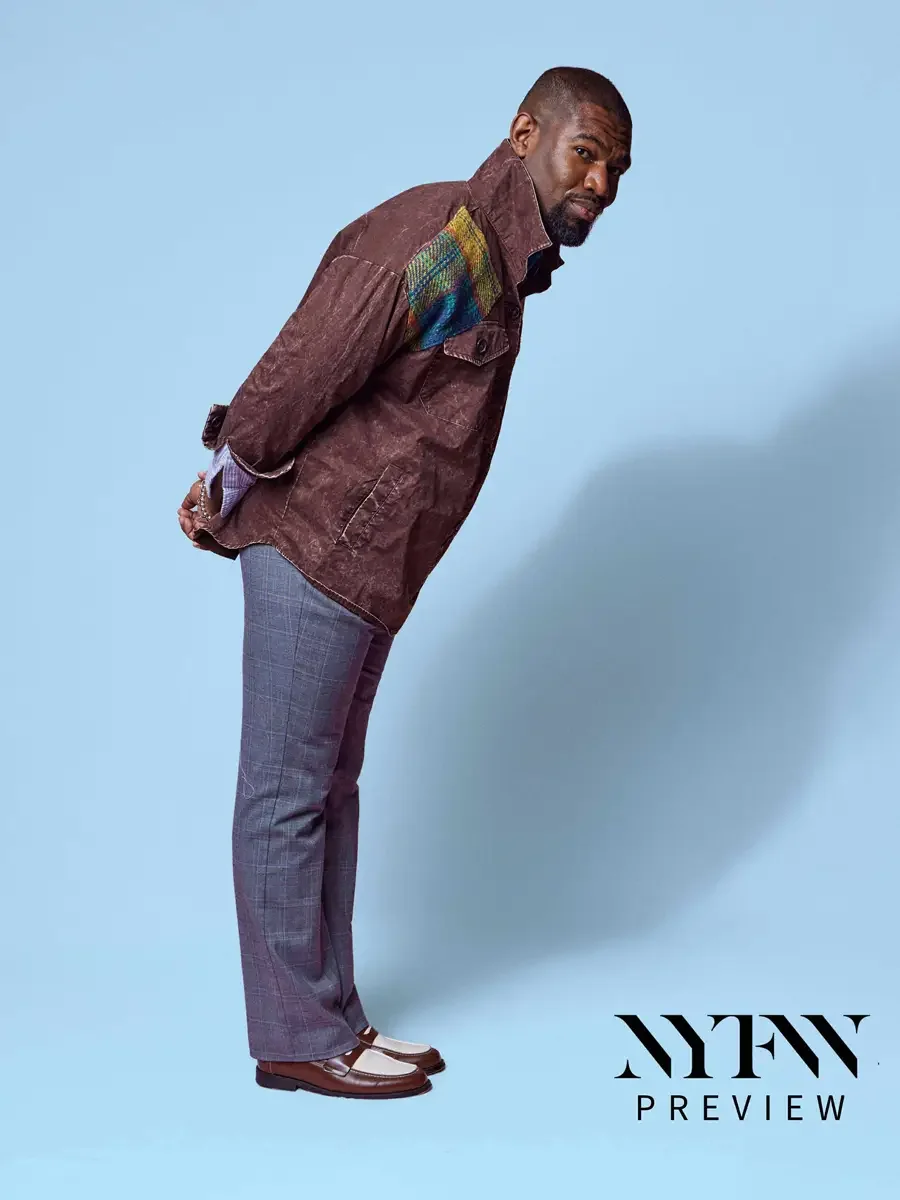 JUS10H 2025 promotional image, model in profile, brown washed field jacket with multicolor tartan patchwork shoulder panel over glen plaid trousers and two-tone loafers, light blue background, photographed by Scott Parker Photo