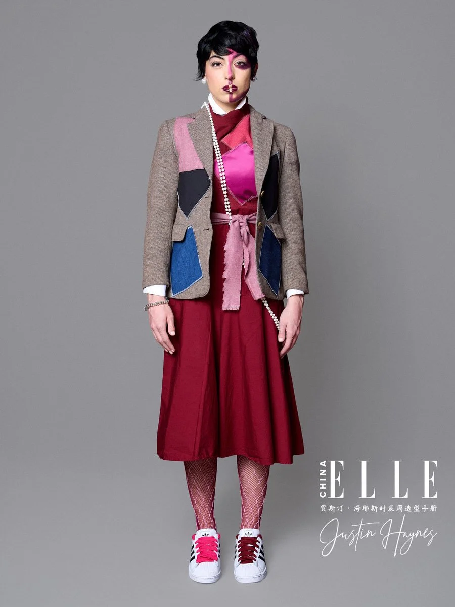 Fashion model standing against a plain gray background, wearing a colorful outfit including a multicolored blazer, pink top, red skirt, patterned tights, and sneakers, with makeup and jewelry, for a fashion magazine photoshoot.
