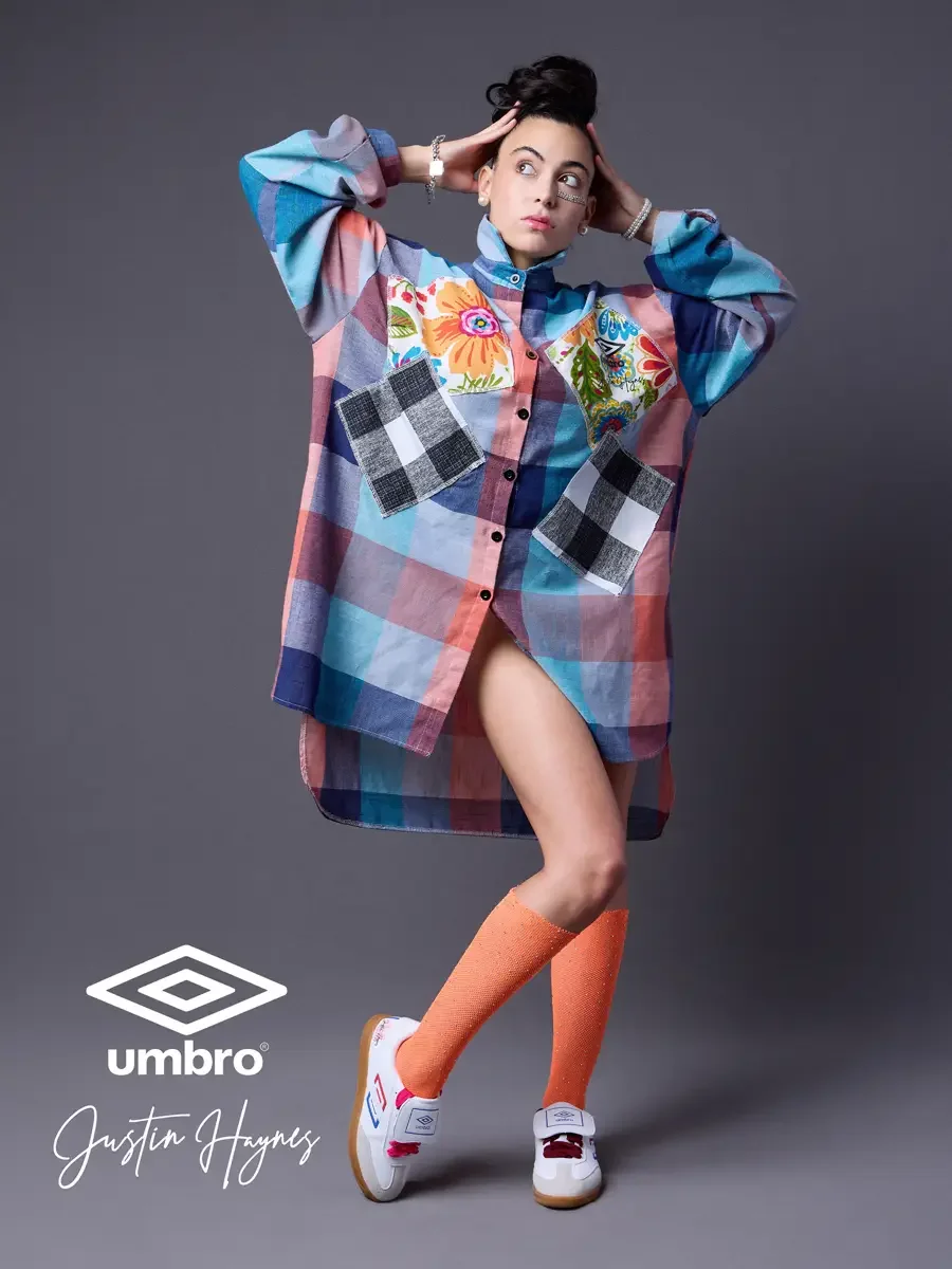 Fashion photographer producer and creative director work for the Umbro France collection. Vanessa in motion with both hands raised to her head and one knee lifted in an oversized multicolor plaid shirt dress with floral patchwork panels and orange fo