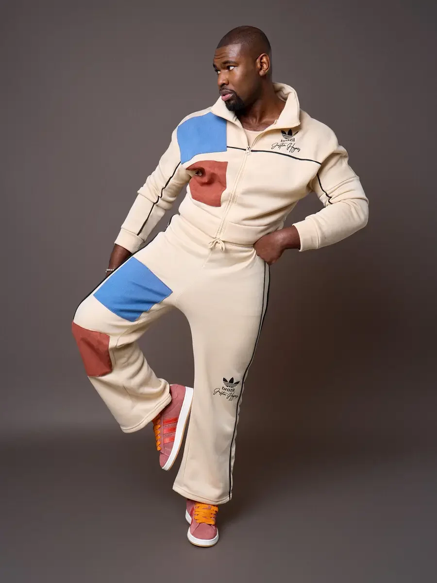JUS10H x Adidas Brasil campaign, model balancing on one leg with knee raised, cream patchwork tracksuit with blue and terracotta suede panels and black piping, Adidas Campus 00s in pink suede with red stripes and orange laces, warm brown background, 