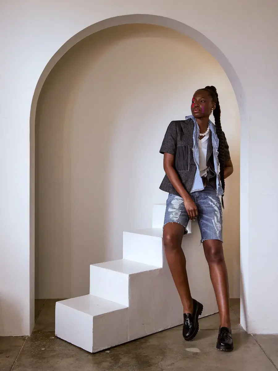 JUS10H Black Summer Jacket content library, female model in dark denim structured vest layered over light blue denim jacket, standing beside white steps inside arch looking up, distressed bermuda shorts, pearl necklace, black loafers, warm neutral ba