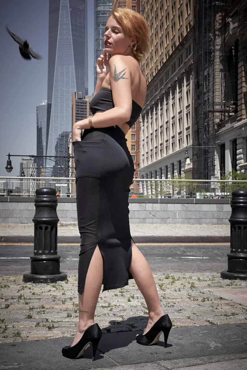 Satine, full length, back to camera, mid-stride, black strapless bandeau crop top, black satin fitted midi skirt with front slit hem and sheer mesh hip panel, black satin peep-toe stiletto heels, gold bangles, bird tattoo on right upper arm visible, 