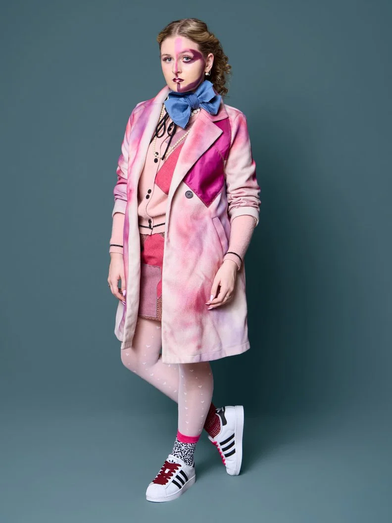 A woman with colorful, artistic makeup and styled hair poses against a teal background. She is dressed in a pink tie-dye coat, pink and red clothing, patterned tights, and white sneakers with black stripes, red laces, and pink accents. She also wears