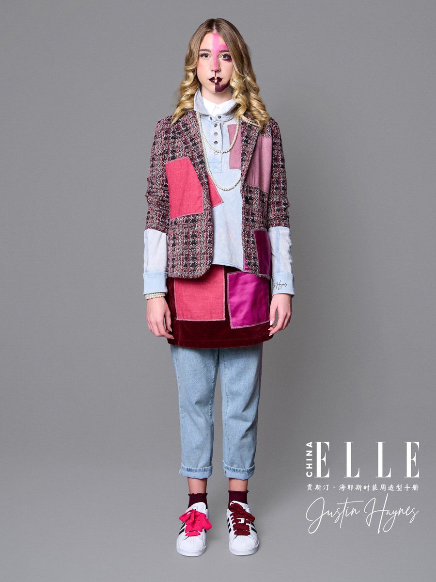 Young woman with blonde wavy hair wearing a colorful patchwork blazer, light denim jeans, and white sneakers with pink laces, standing against a gray background.