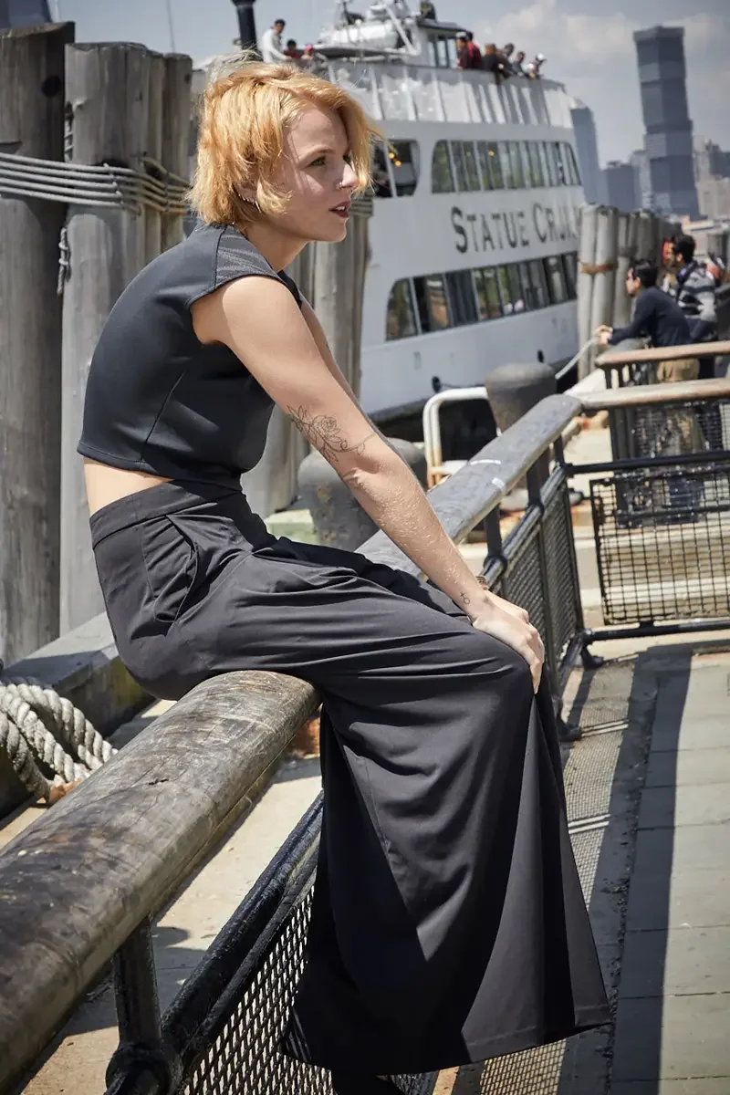 Satine, full length, seated on pier railing, profile, black structured cap-sleeve crop top, black wide-leg pleated palazzo pants, floral tattoo on left forearm visible, Statue Cruises ferry in background, Battery Park pier test shoot, photographed by