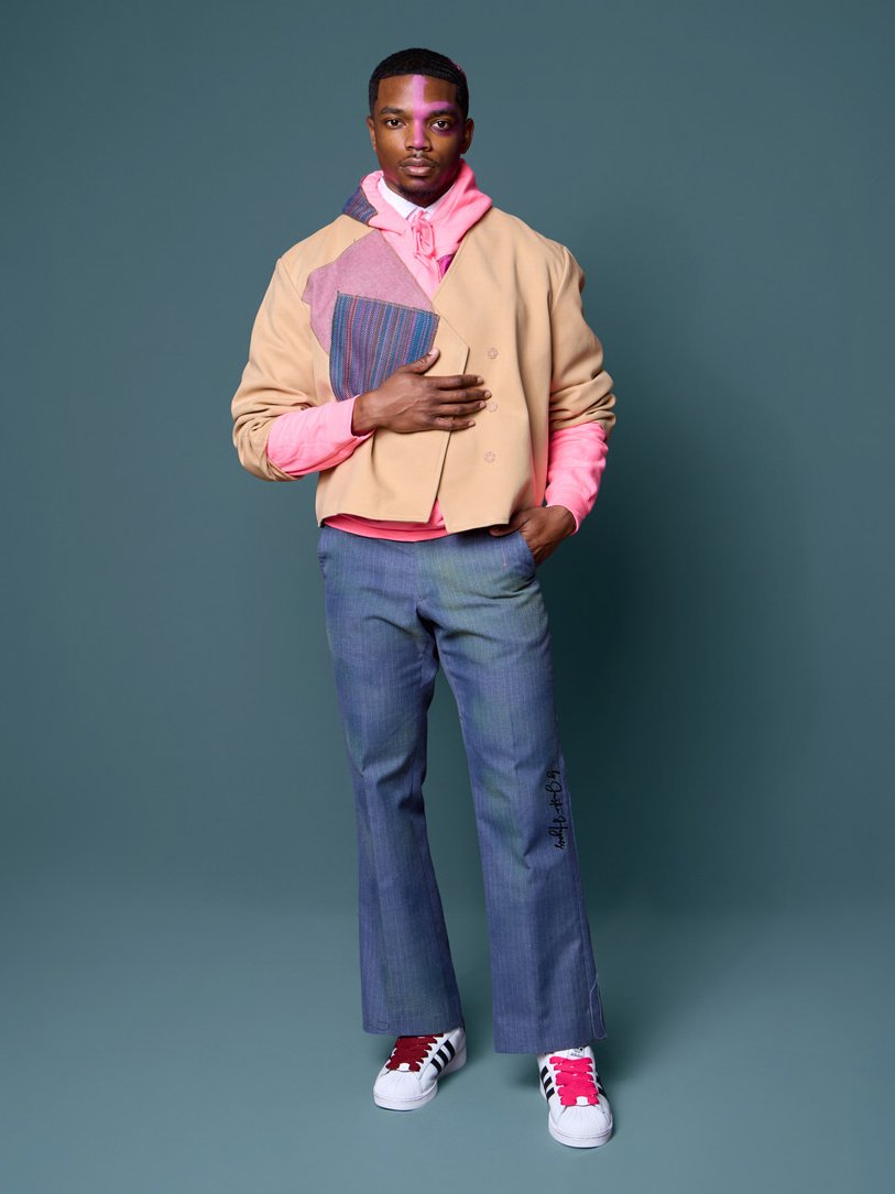 A young man standing against a teal background, dressed in a colorful, layered outfit that includes a beige jacket with patchwork details, pink hoodie, gray plaid pants, and white sneakers with red and black accents.