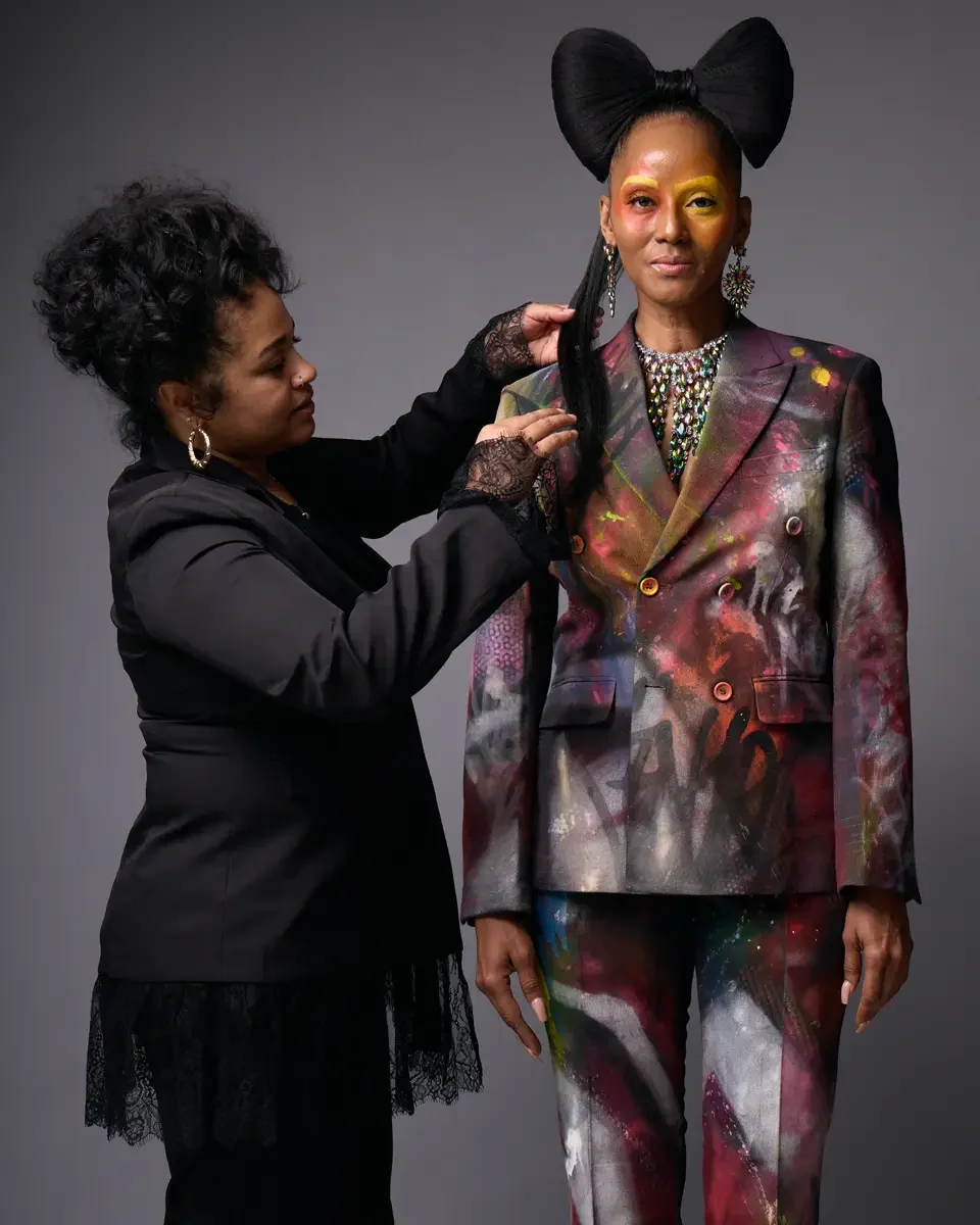 JUS10H Met Monday 2026, hairstylist adjusting model's hair on set, hand-painted suit with iridescent necklace, grey background, photographed by Scott Parker Photo