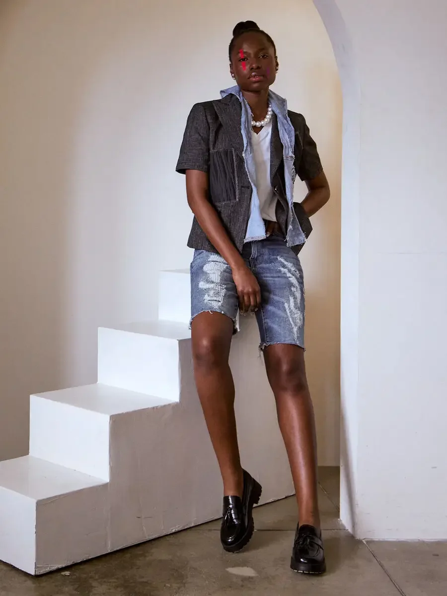 JUS10H Black Summer Jacket content library, female model in dark denim structured vest layered over light blue denim jacket, distressed bermuda shorts, pearl necklace, black loafers, leaning against white steps inside arch, warm neutral background, p